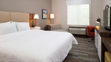 Quarto de hotel com uma cama grande, uma escrivaninha com computador, uma cadeira, uma televisão e uma janela com uma cobertura vermelha.