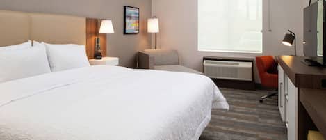 Habitación de hotel con una cama grande, un escritorio con computadora, una silla, un televisor y una ventana con cortinilla roja.