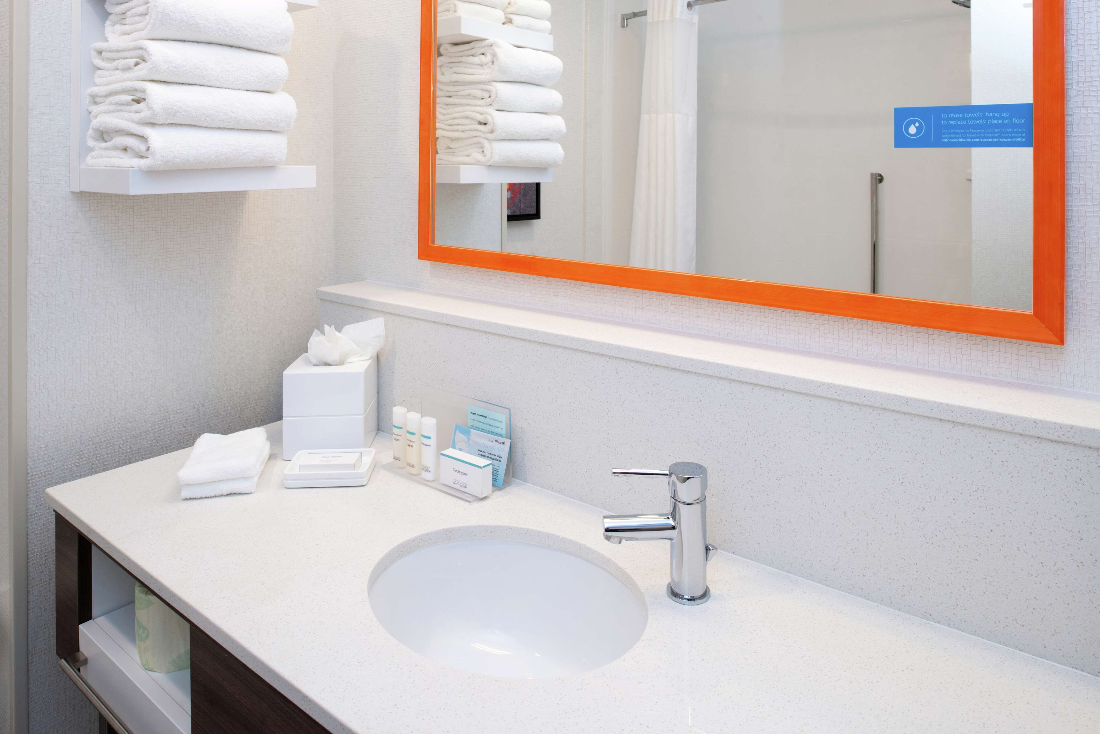 A modern bathroom with a white sink, a mirror with an orange frame, and neatly folded towels on a shelf.