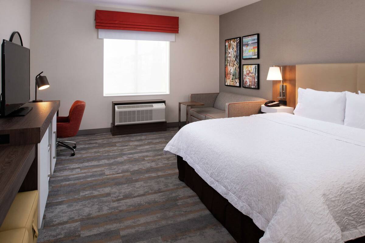 Une chambre d’hôtel avec un grand lit, un bureau avec un ordinateur, une chaise, un canapé, un radiateur et des œuvres d’art accrochées au mur.