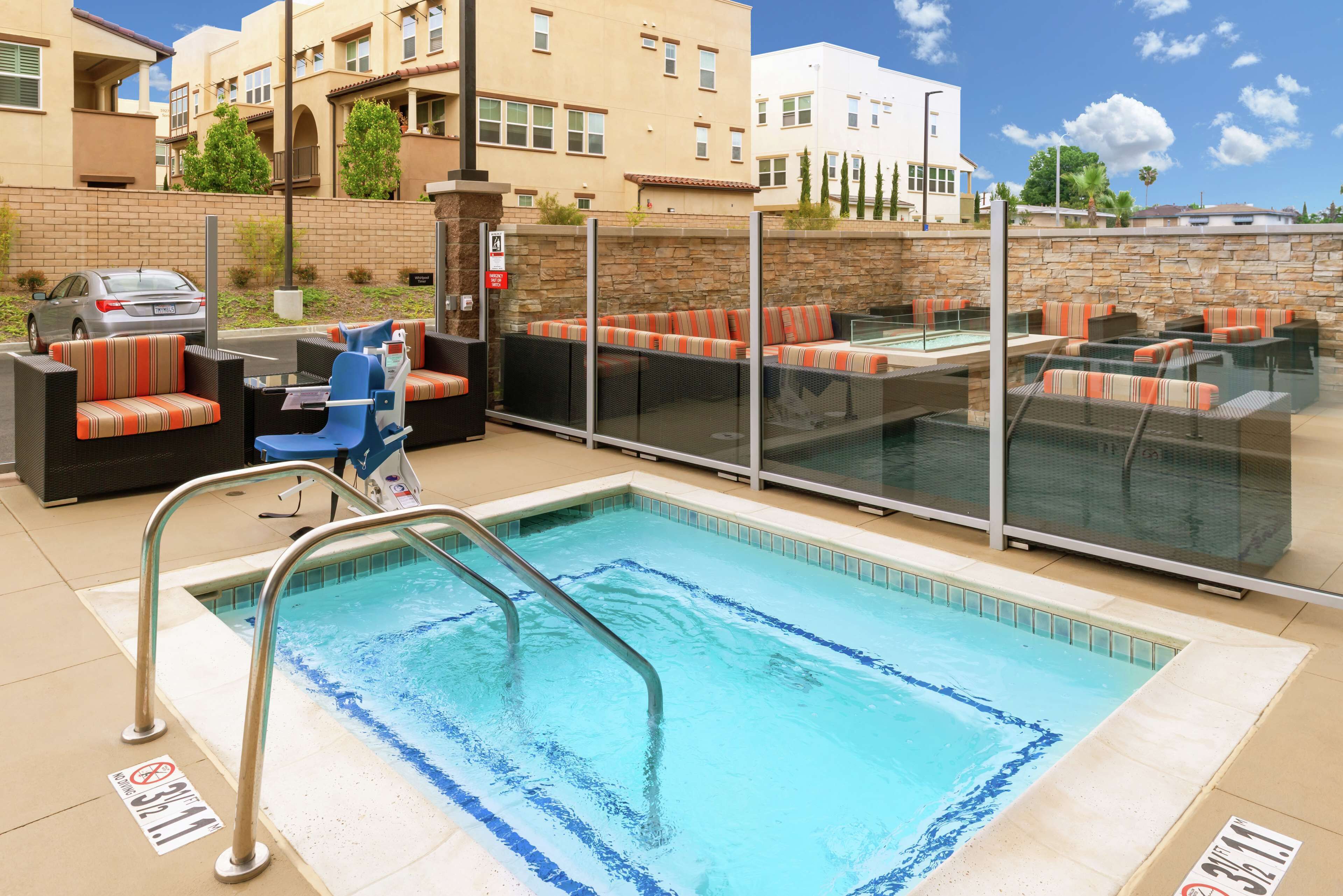 Una piscina con mobiliario de descanso y una zona cubierta, rodeada por un muro de piedra y edificios residenciales.