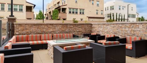 An outdoor patio area with wicker furniture and a glass-top table, surrounded by stone walls and modern buildings in the background.