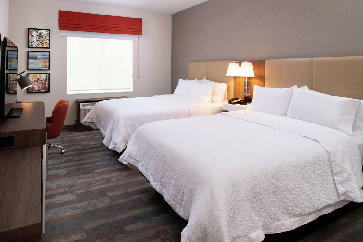 Une chambre d’hôtel avec deux lits, un bureau, une chaise, une lampe et des œuvres d’art accrochées au mur.