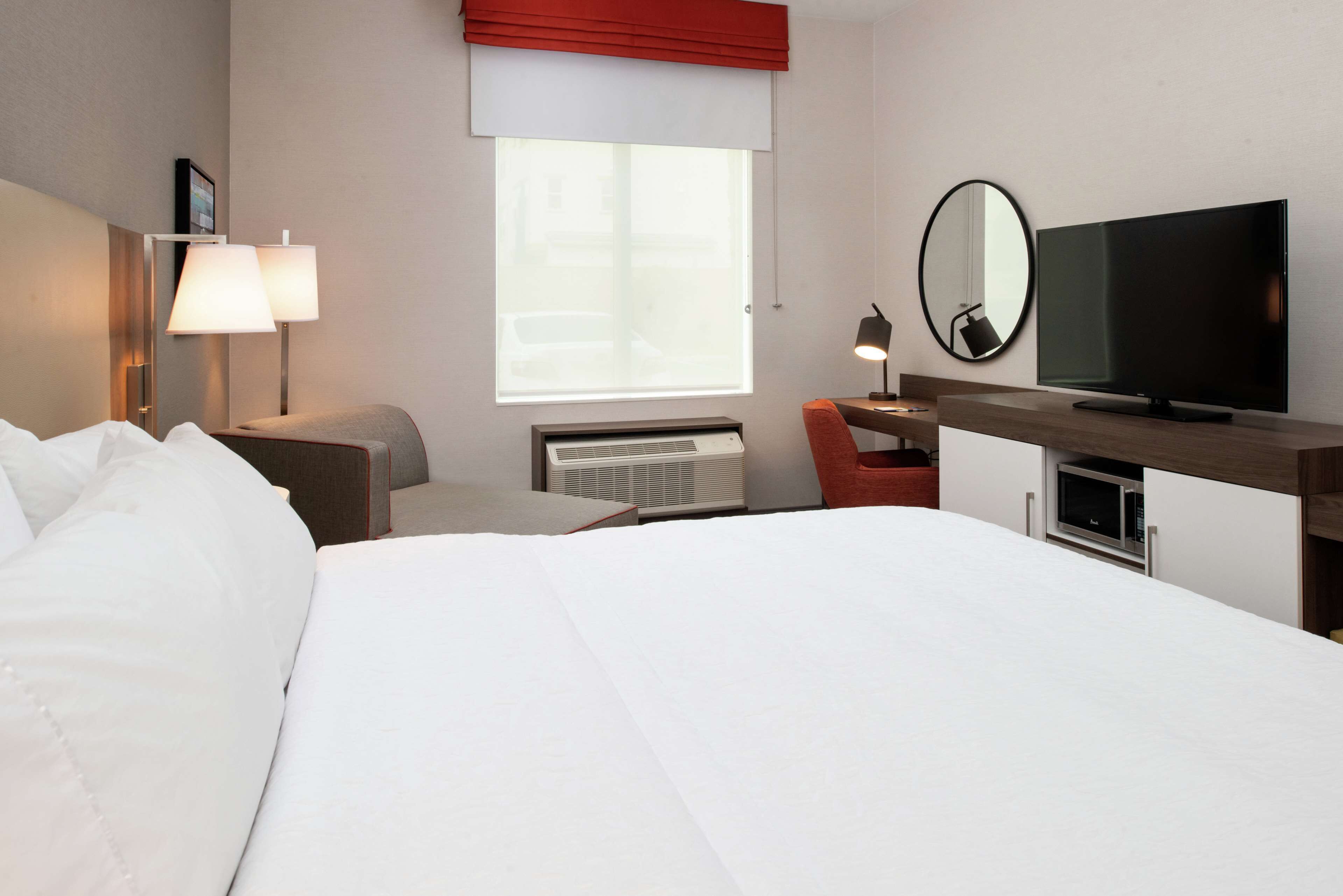 A hotel room with a bed, a desk with a computer, a television, a chair, and a window with a view.