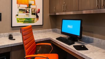 A hotel room with a desk, a computer, an orange chair, a framed picture on the wall, and wooden cabinets.