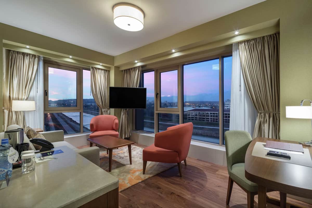 A hotel room with a large window, a flat-screen TV, two armchairs, a small table, and a desk with a lamp.