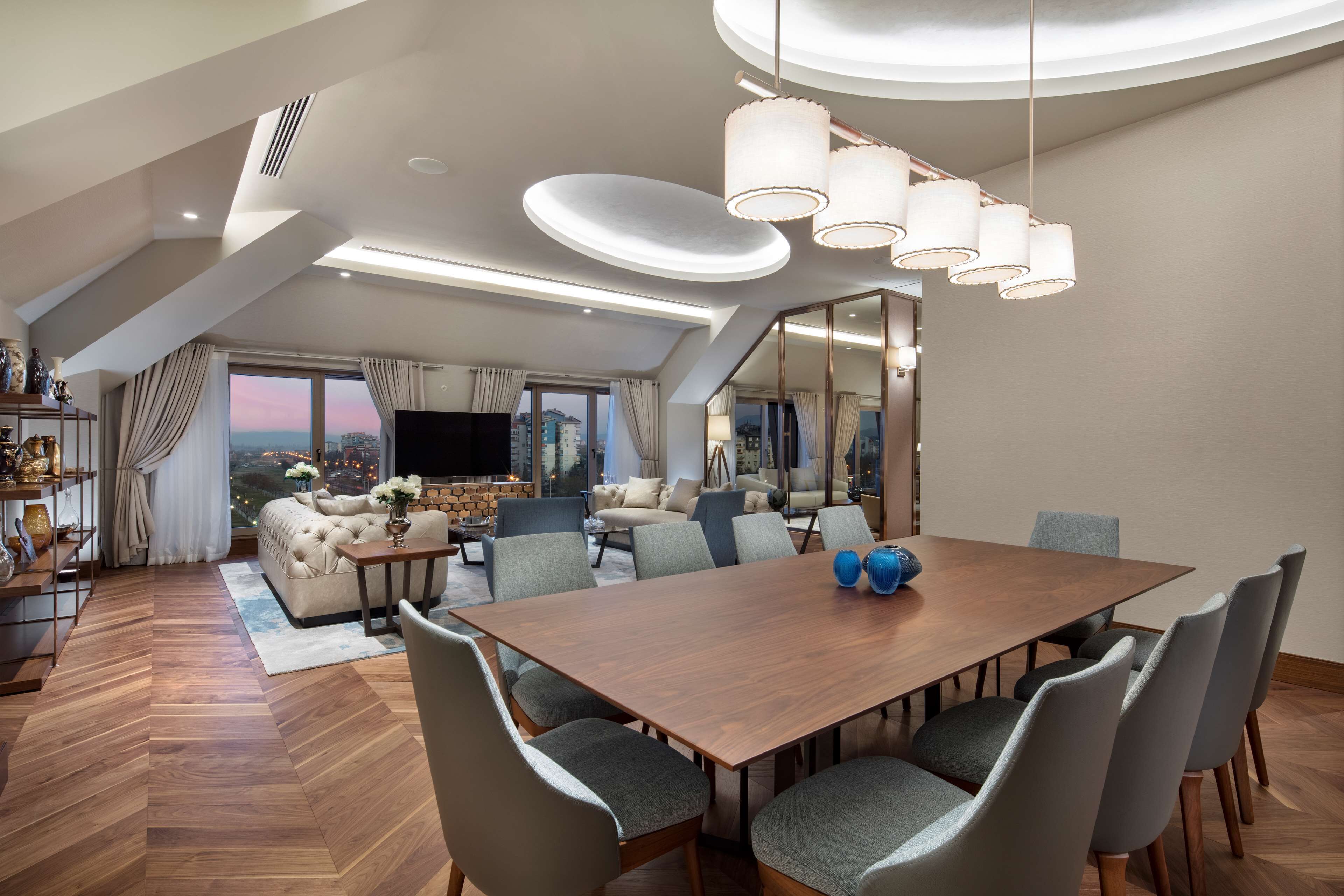 A spacious living area with a large wooden dining table, grey upholstered chairs, and a modern chandelier.