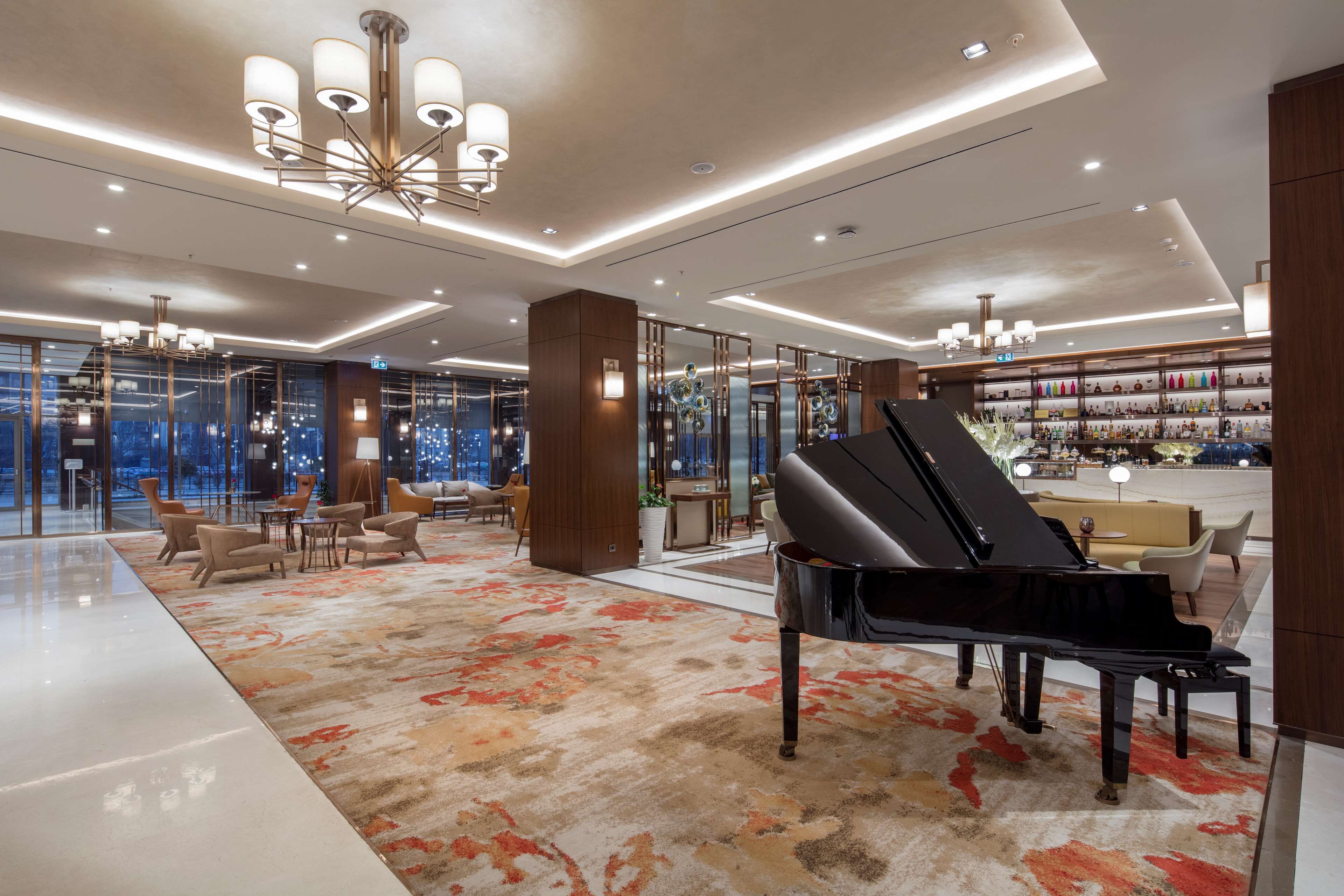 Um hall amplo com um piano de cauda, luminárias modernas e um bar com diversas garrafas.