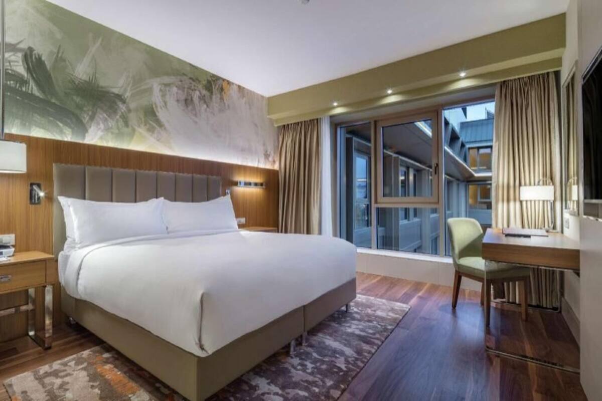 A hotel room with a large bed, a desk, a chair, a TV, and a large window with curtains.