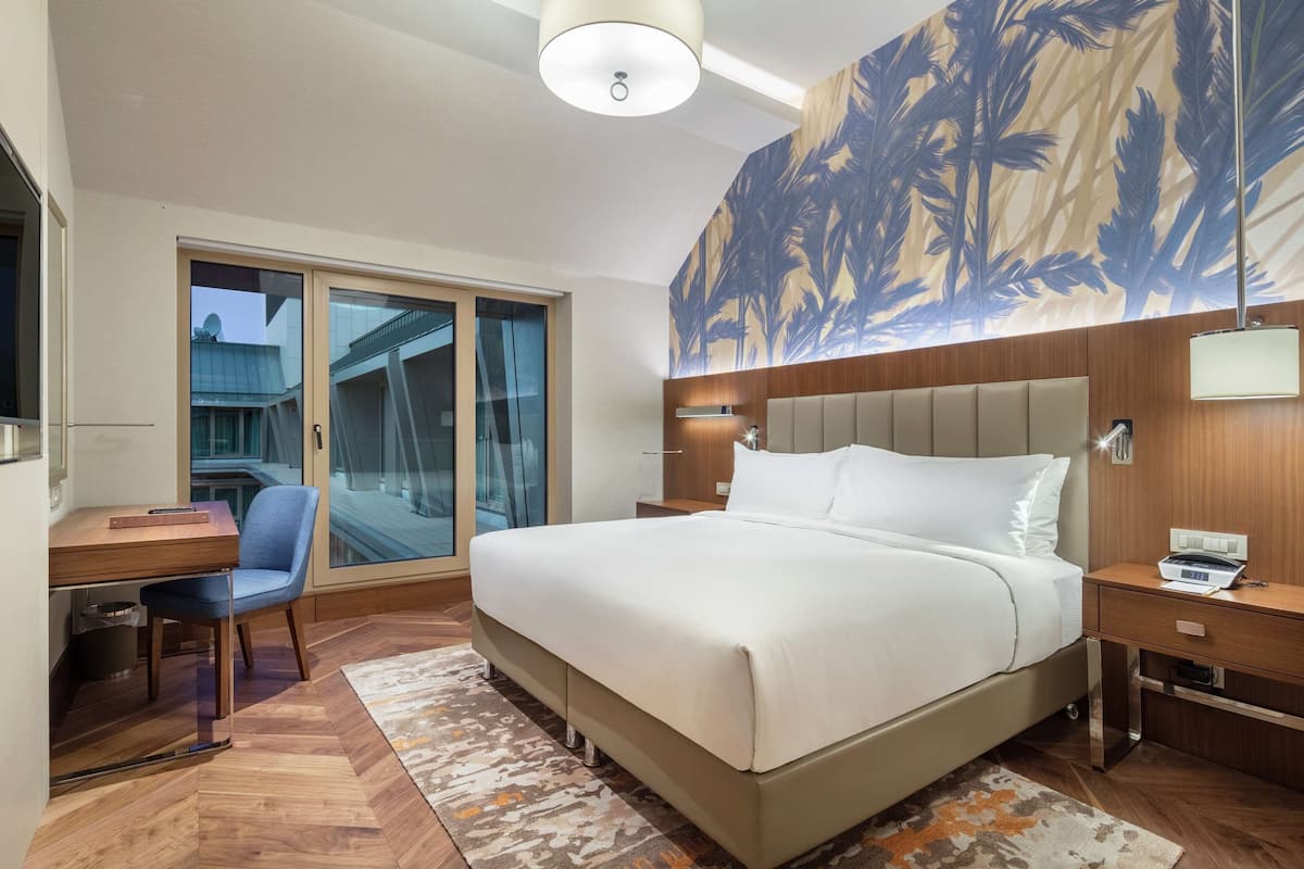 A modern hotel room with a large bed, a desk with a chair, a TV, and a view of the outside.