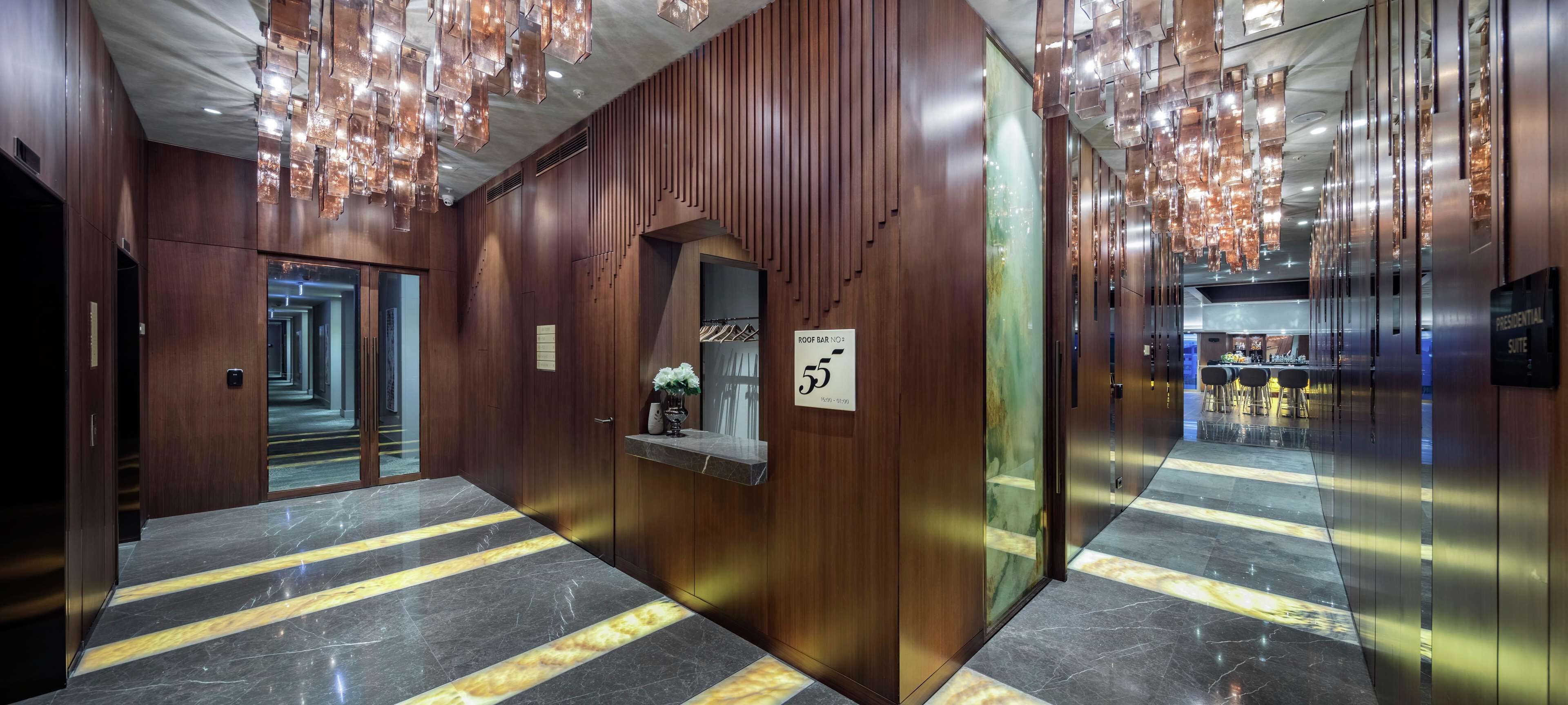 A modern hotel corridor with wooden paneling, a marble floor with yellow stripes, and a ceiling with hanging lights.