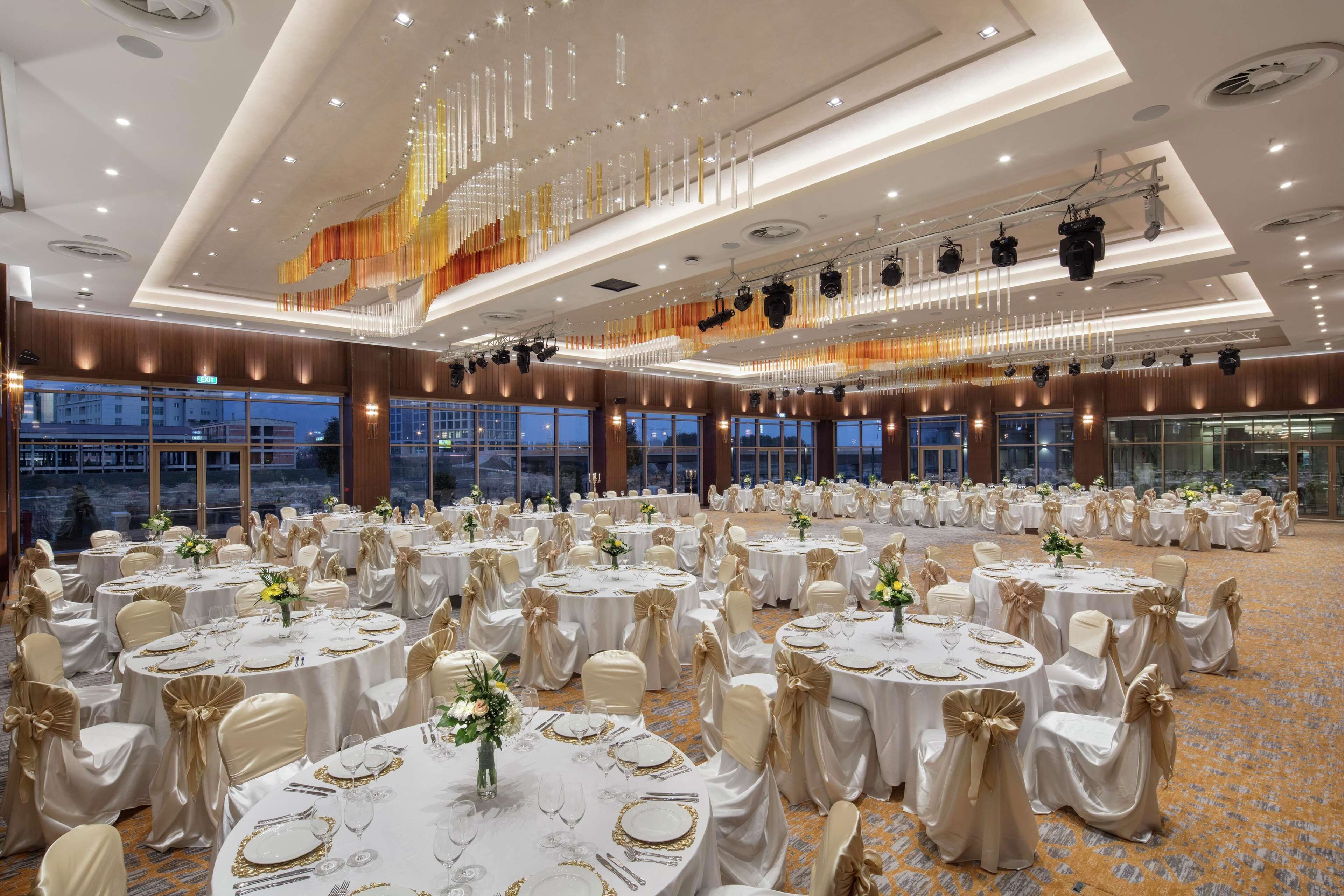 Une salle de banquet avec des tables rondes disposées pour les événements, de grandes fenêtres et un design de plafond moderne.