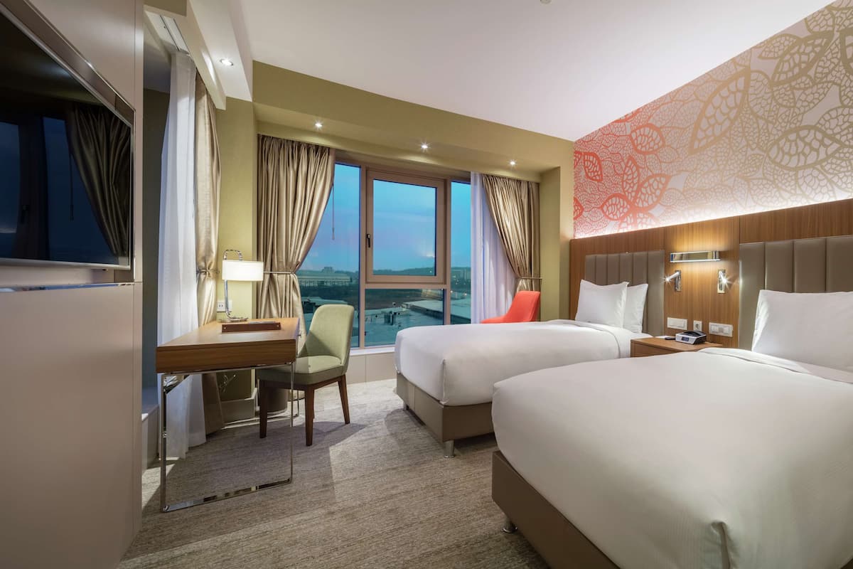 A hotel room with two beds, a desk, a chair, and a large window with a view.