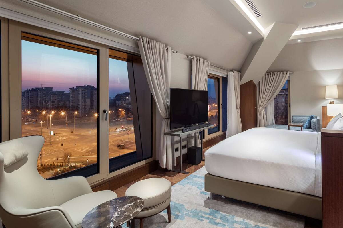 A hotel room with a large bed, a TV, a desk, and a view of a cityscape at dusk.