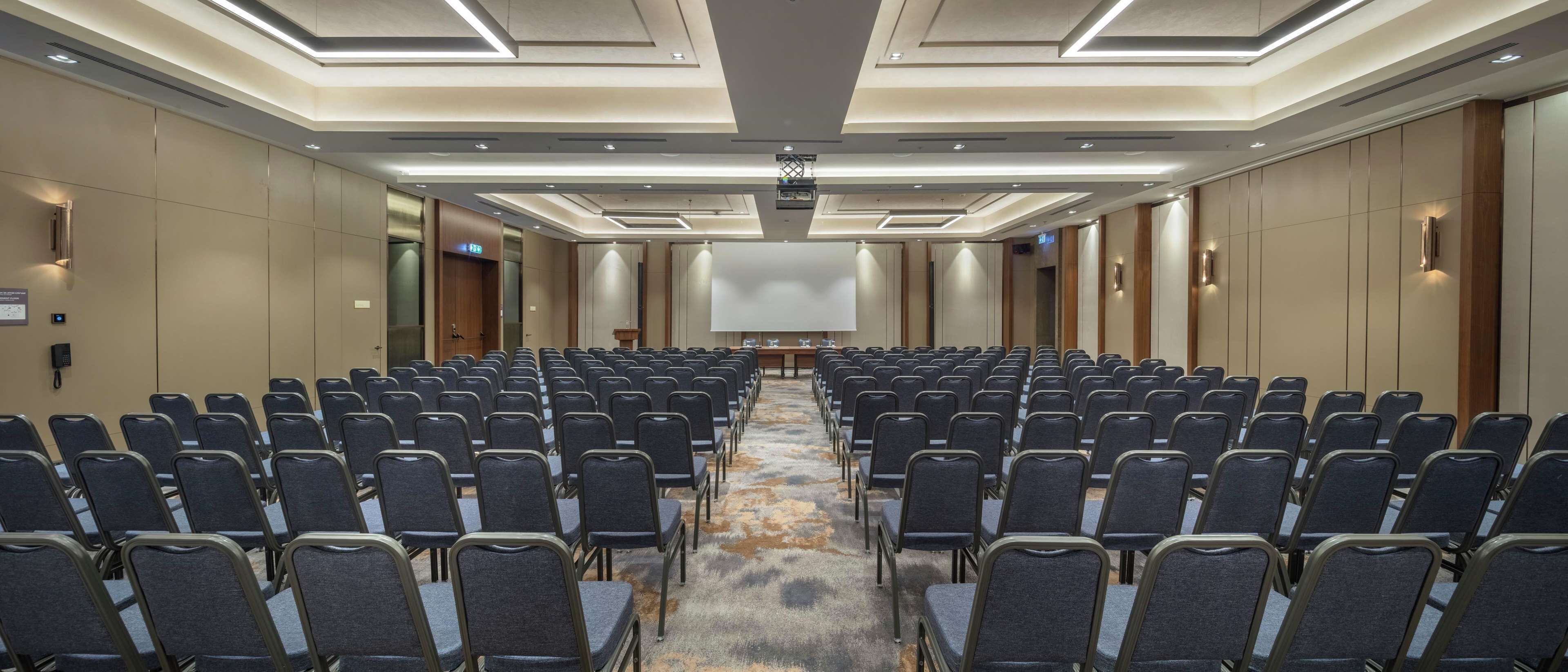 Une salle de conférence avec des rangées de chaises, un grand écran et un sol recouvert de moquette.