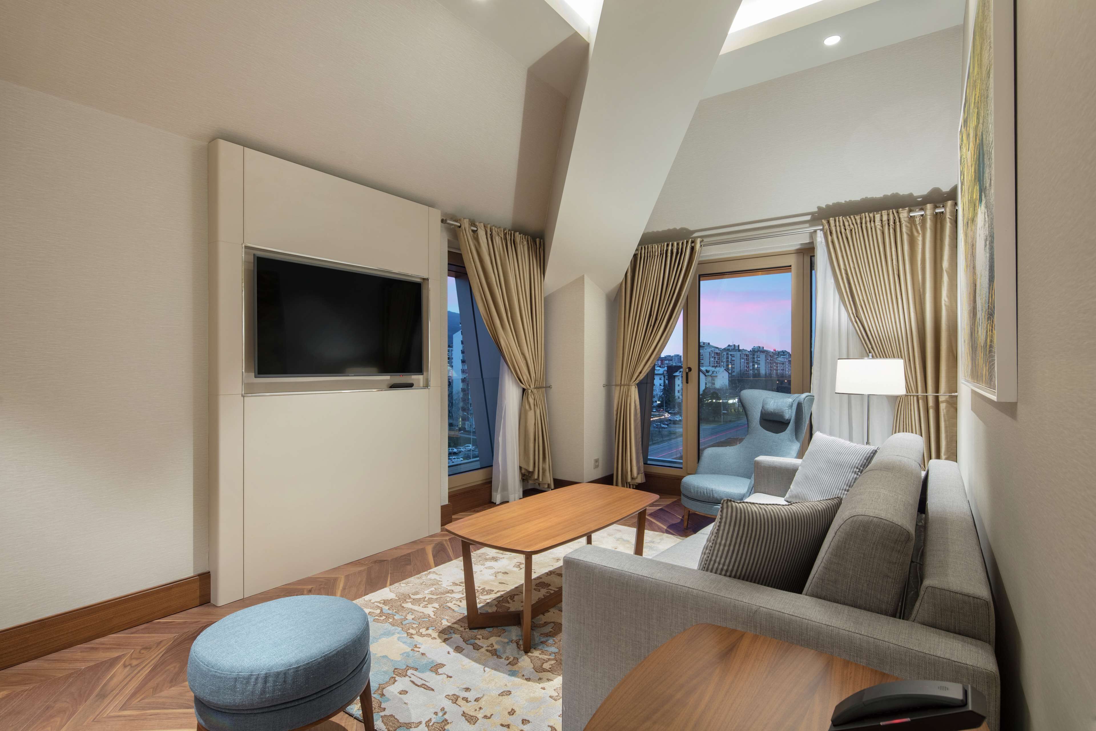 A modern hotel room with a sofa, a small table, a TV, and a view of the city through large windows.