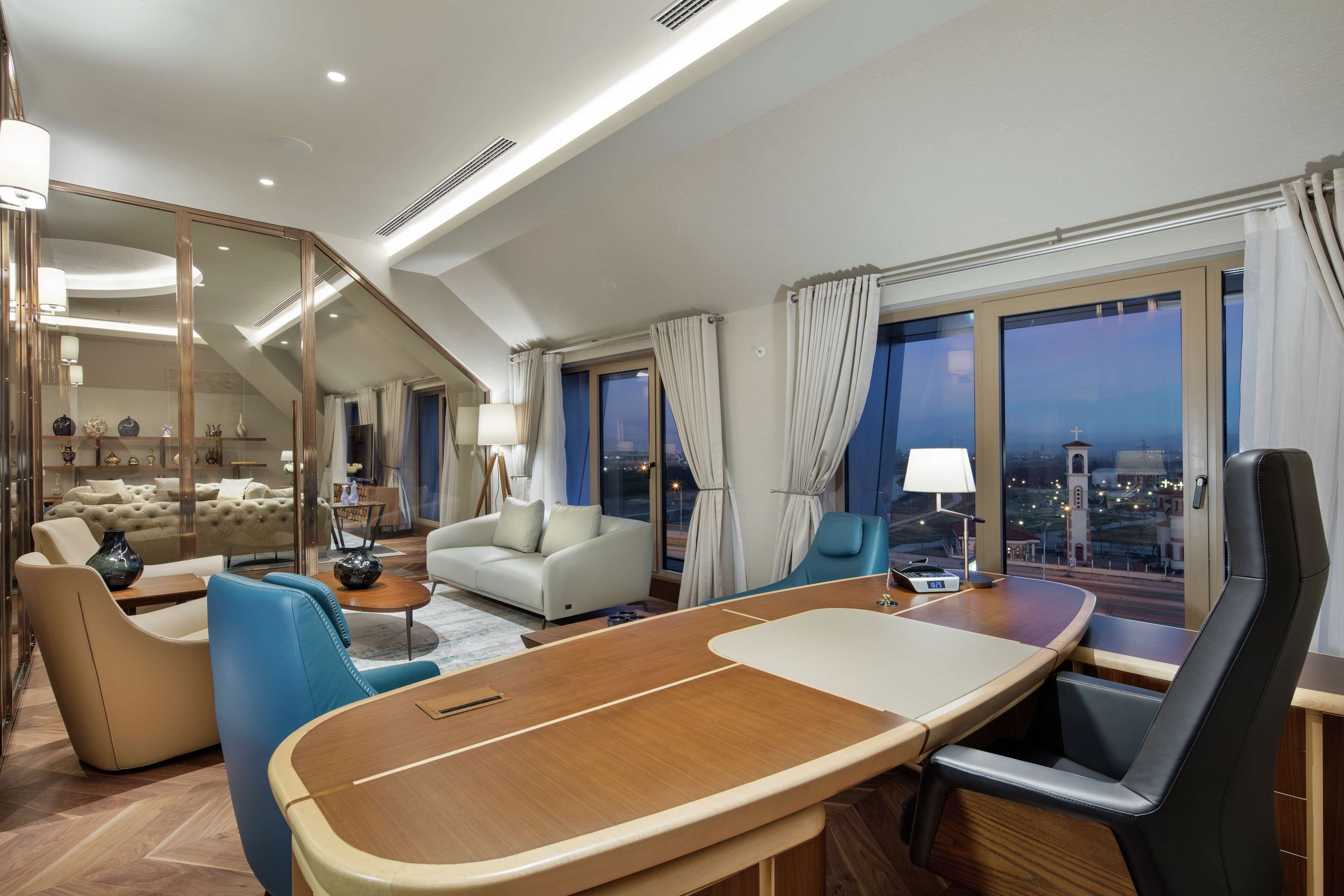 A modern hotel room with a curved wooden desk, a sofa, a chair, and a view of the cityscape.