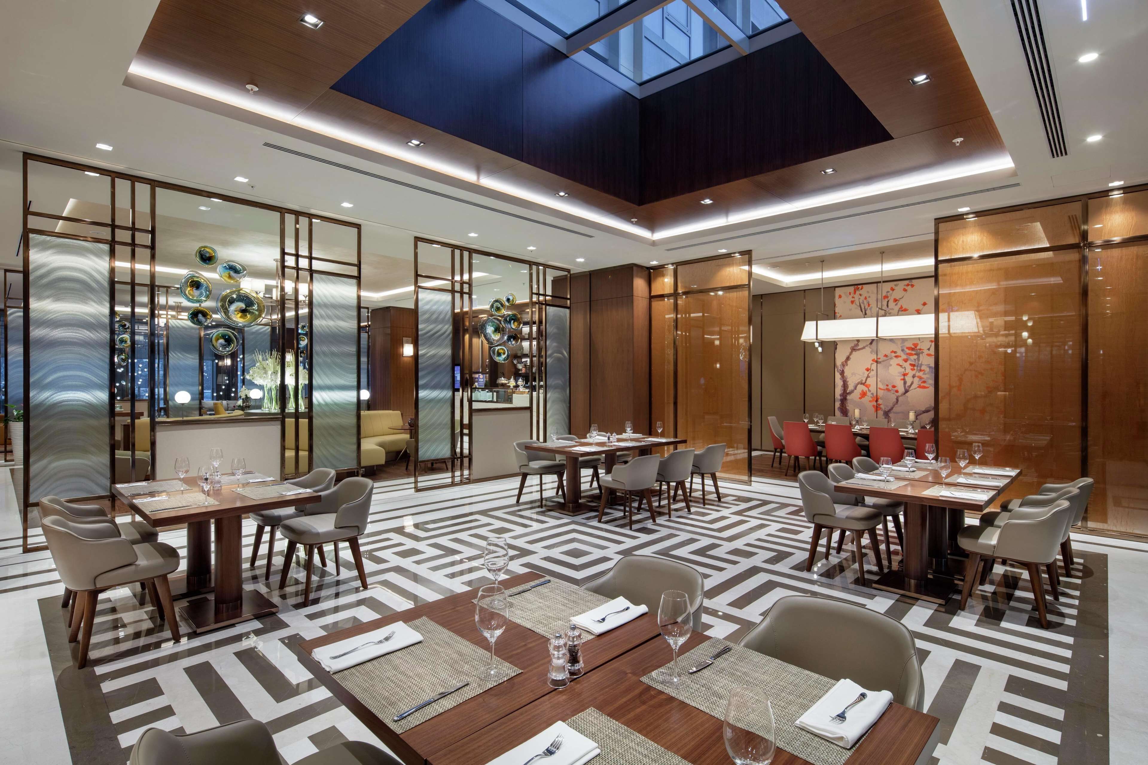 Área de jantar de um hotel moderno, com várias mesas preparadas para refeições, teto alto com claraboias e painéis de vidro decorativos.