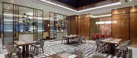 Área de jantar de um hotel moderno, com várias mesas preparadas para refeições, teto alto com claraboias e painéis de vidro decorativos.
