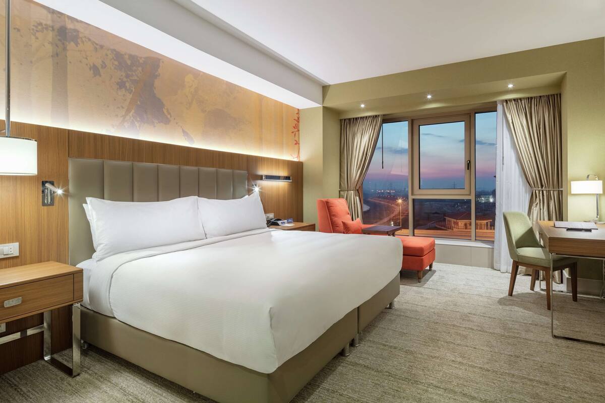 A hotel room with a large bed, a desk, a chair, and a view of the city at sunset.