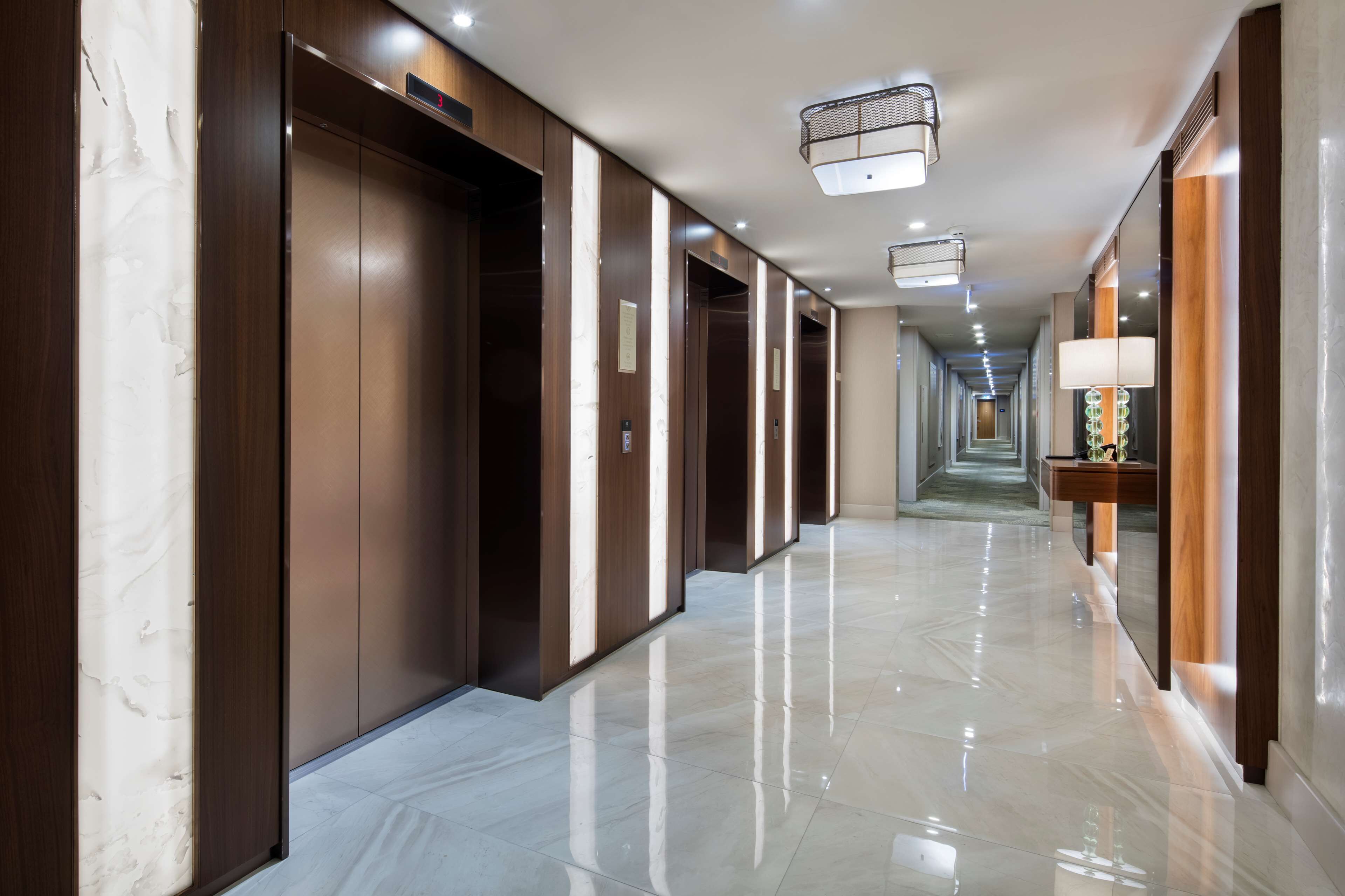 A long, polished hallway with wooden doors and a marble floor, leading to a distant elevator.