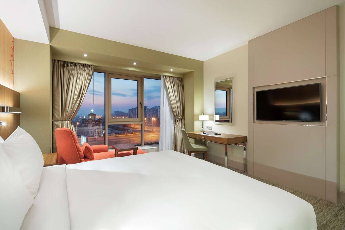 A hotel room with a large bed, a desk, a chair, a TV, and a view of the city at dusk.