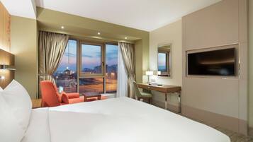 Quarto de hotel com uma cama grande, uma mesa, uma cadeira, uma televisão e vista para a cidade ao entardecer.