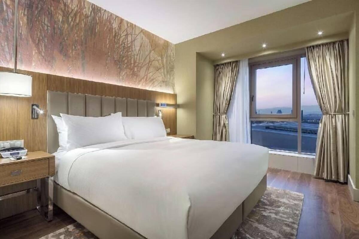 A hotel room with a large bed, a nightstand, a lamp, and a window with curtains.