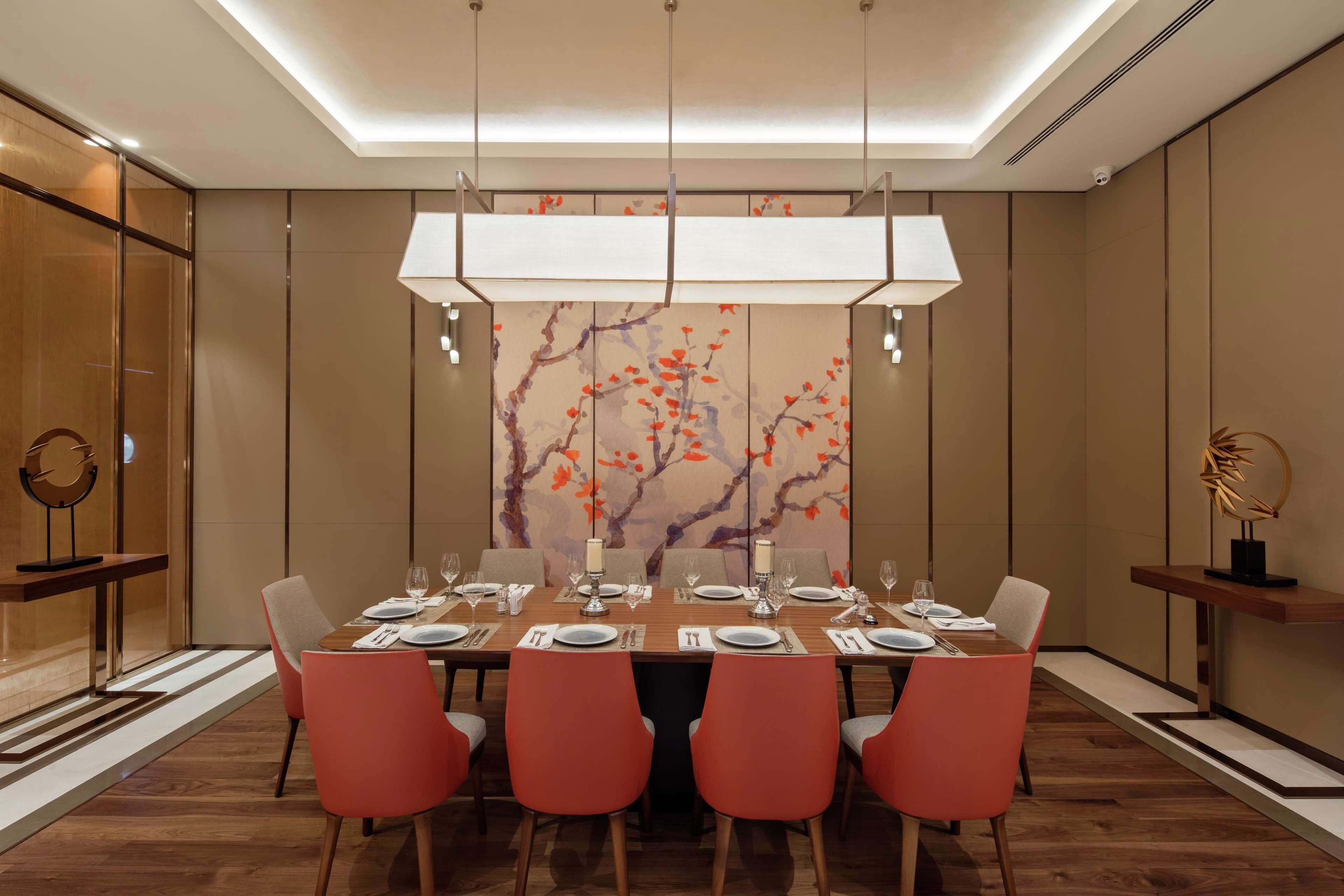 A modern dining room with a long table set for six, featuring red chairs, a decorative wall with a floral pattern, and a ceiling-mounted light fixture.