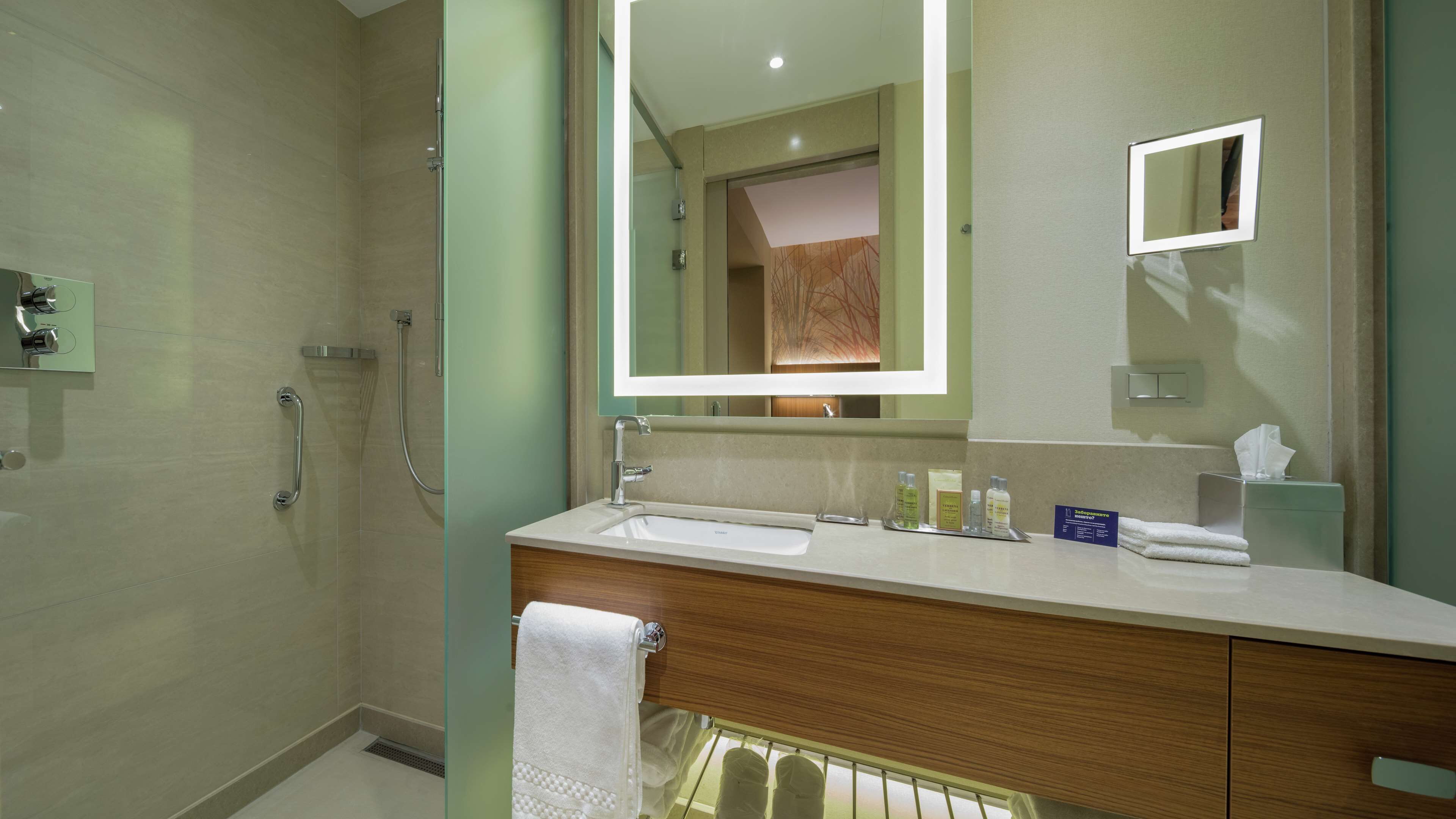 A modern bathroom with a glass shower enclosure, a large mirror, a sink with toiletries, and a towel rack.