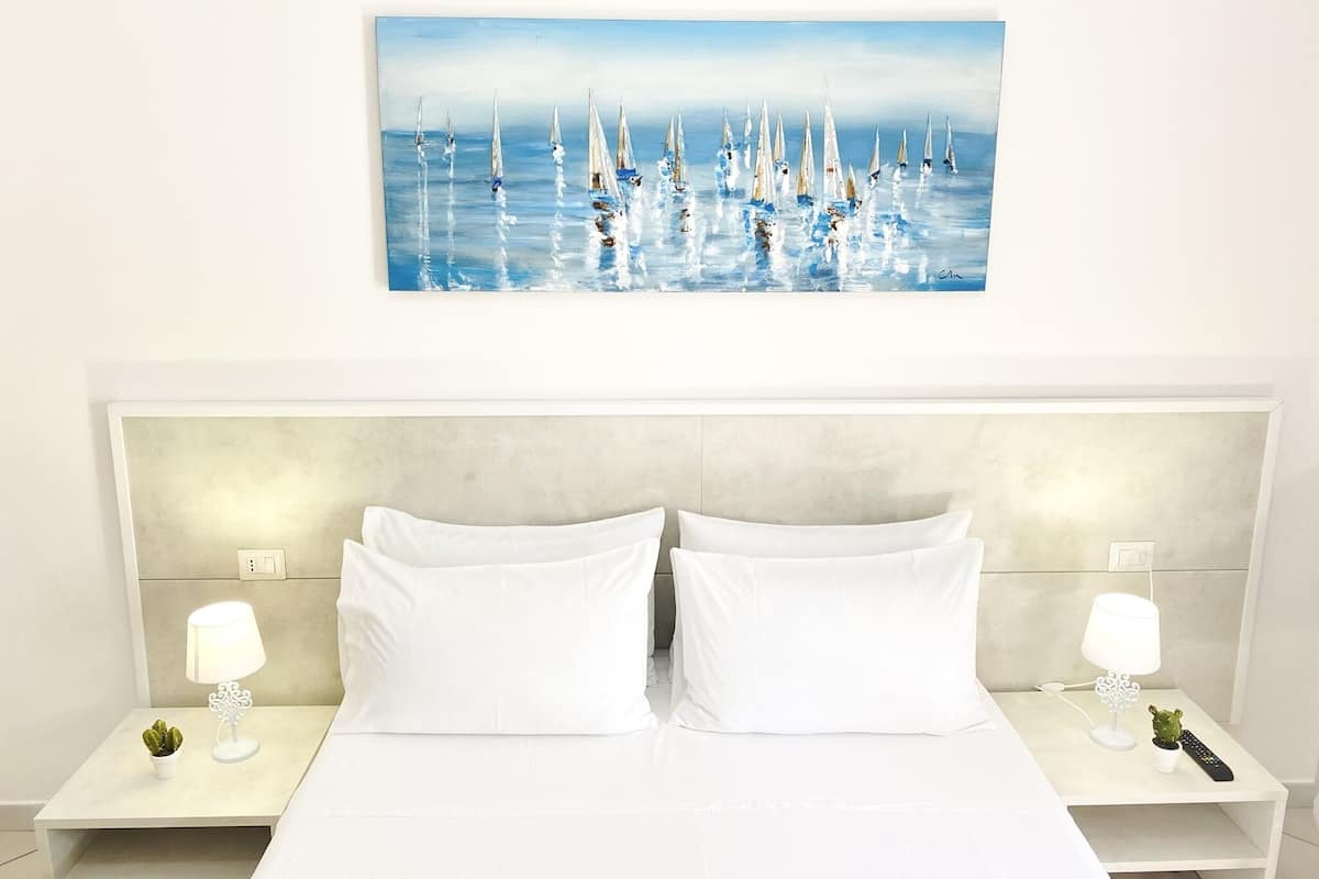 A hotel room with a large bed, white bedding, two bedside tables with lamps, and a painting of sailboats on the wall.
