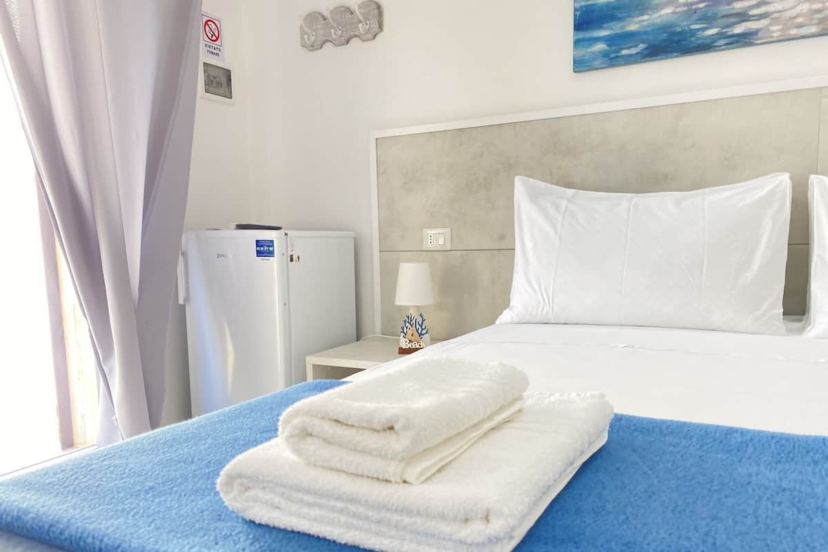 A hotel room with a bed, a small table with a lamp, a refrigerator, and a painting on the wall.