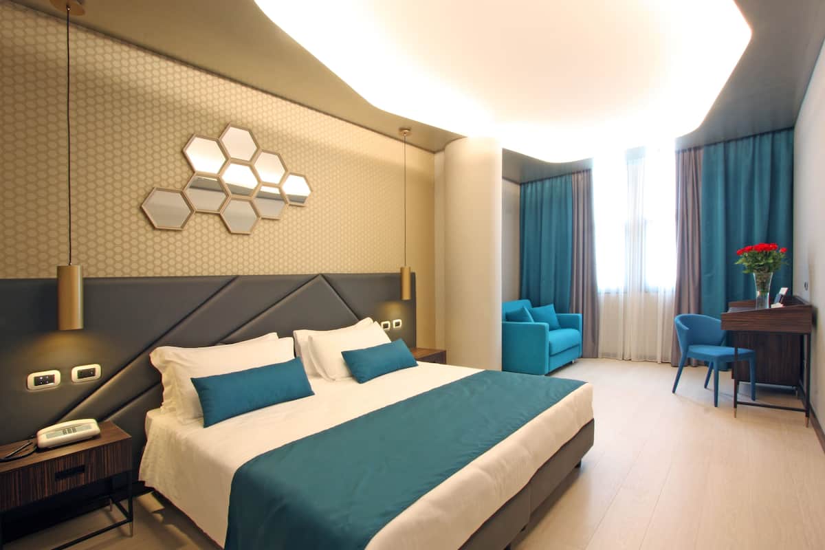 A modern hotel room with a large bed, a desk with a chair, and a seating area with sofas.