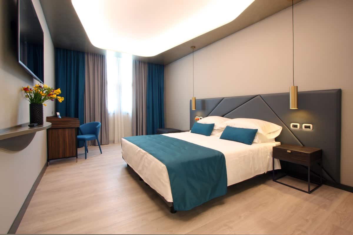 A modern hotel room with a large bed, a desk with a chair, a flat-screen TV, and a vase with flowers.