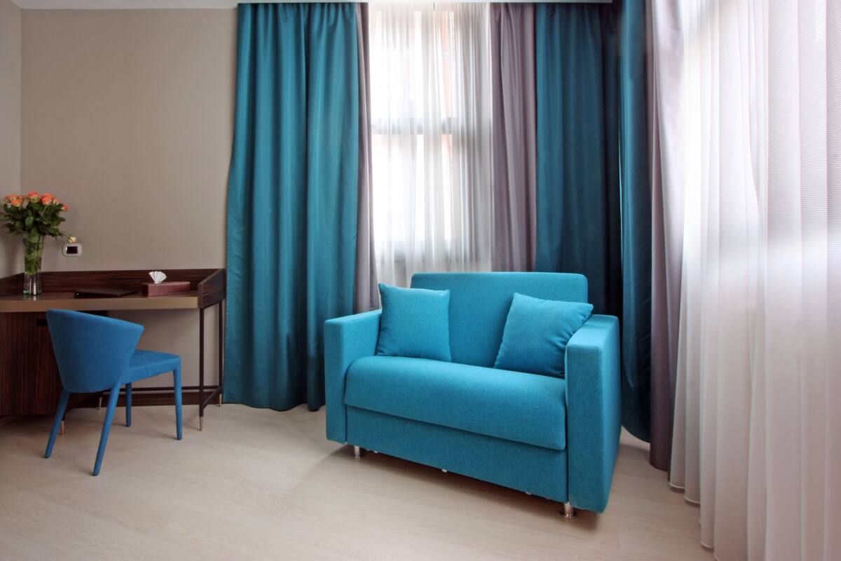 A modern living room with a teal sofa, a blue chair, and a desk with a vase of flowers.