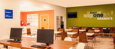 A conference room with two computer stations, white chairs, and a large screen displaying a logo.