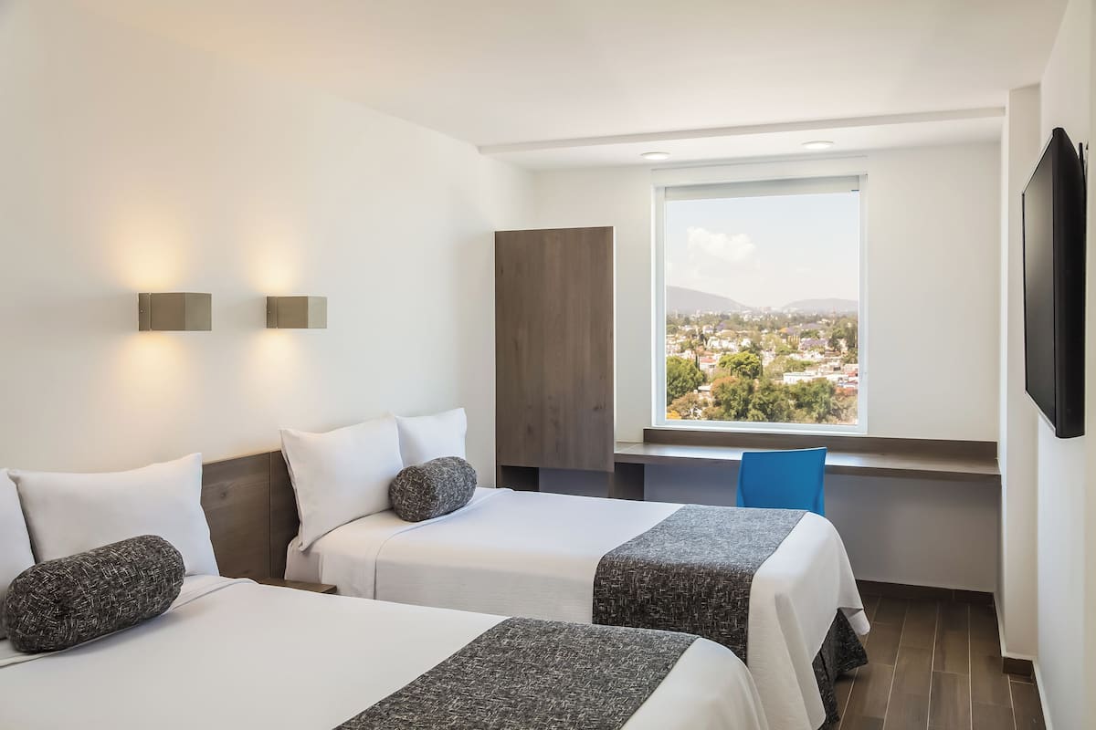 A hotel room with two beds, a large window with a city view, a flat-screen TV, and a blue chair.