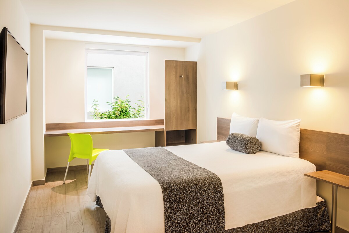 A hotel room with a bed, a desk, a chair, a window, and a wardrobe.