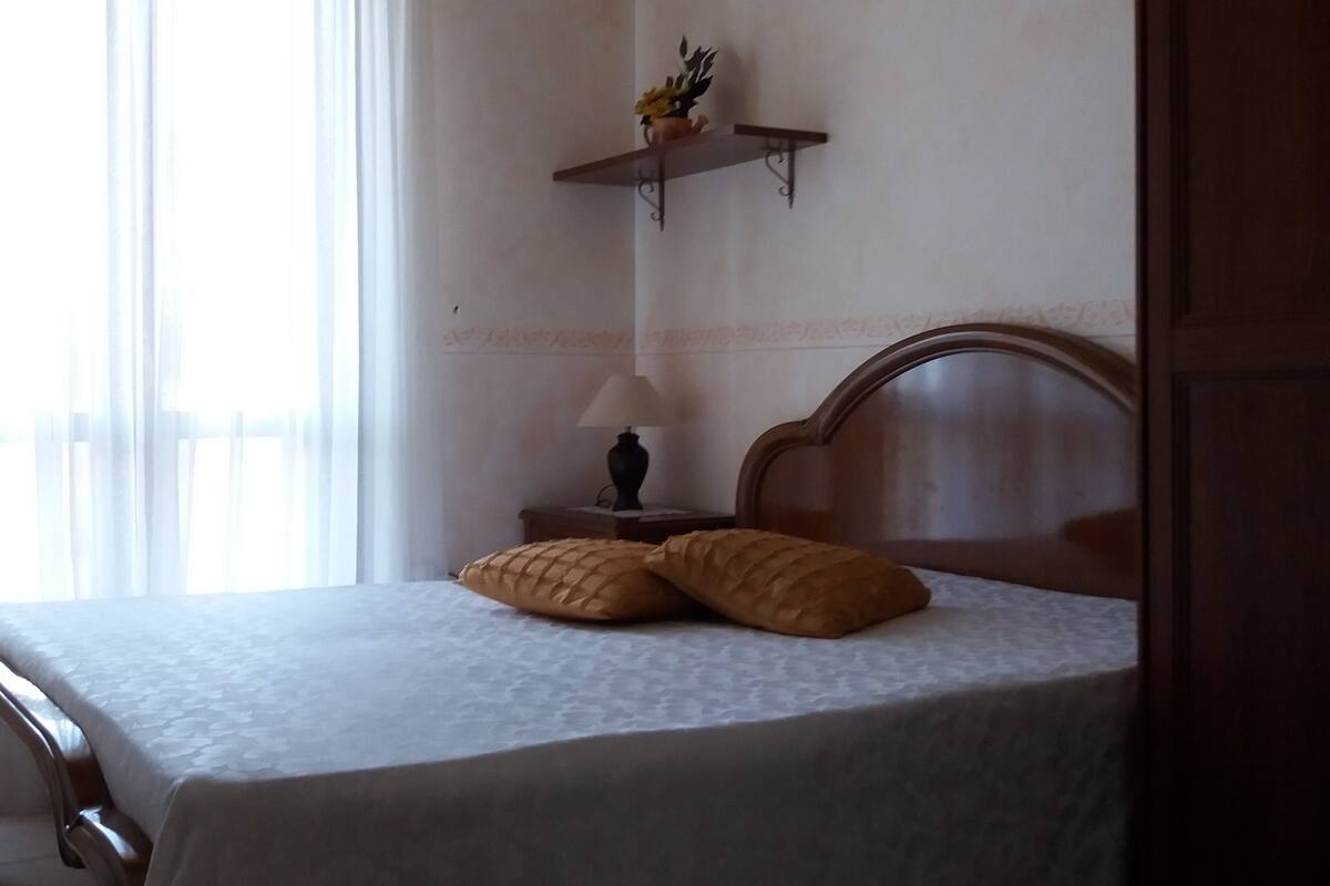Una cama bien hecha con cabecera, una lámpara de noche y un pequeño estante con una flor.