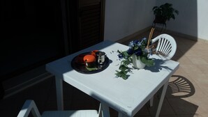 Una pequeña mesa exterior con mantel blanco, una planta en maceta y una bandeja con tazas y un tazón.