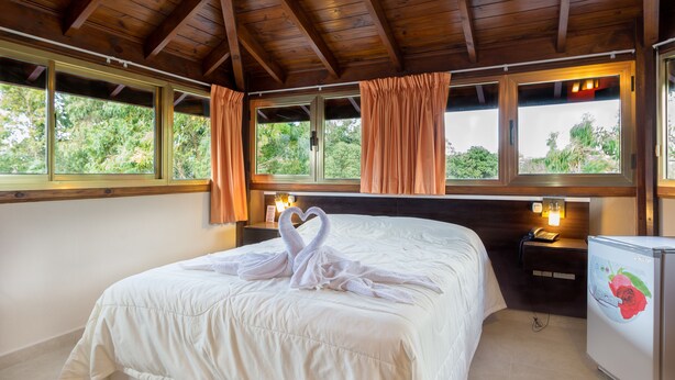 Habitación de hotel con una cama grande, vigas de madera en el techo, ventanas con cortinas, una mesita de noche con lámpara y un refrigerador con una calcomanía de rosa.