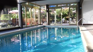 Una piscina interior con paredes de vidrio transparente, borde de azulejos y zona de la piscina con sillas y sombrillas.