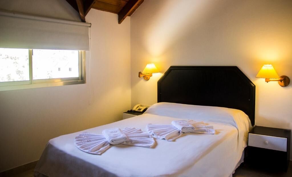 Hotellihuone, jossa on sänky, kaksi yöpöytävalaisinta, yöpöytä ja ikkuna, jossa on sälekaihtimet.