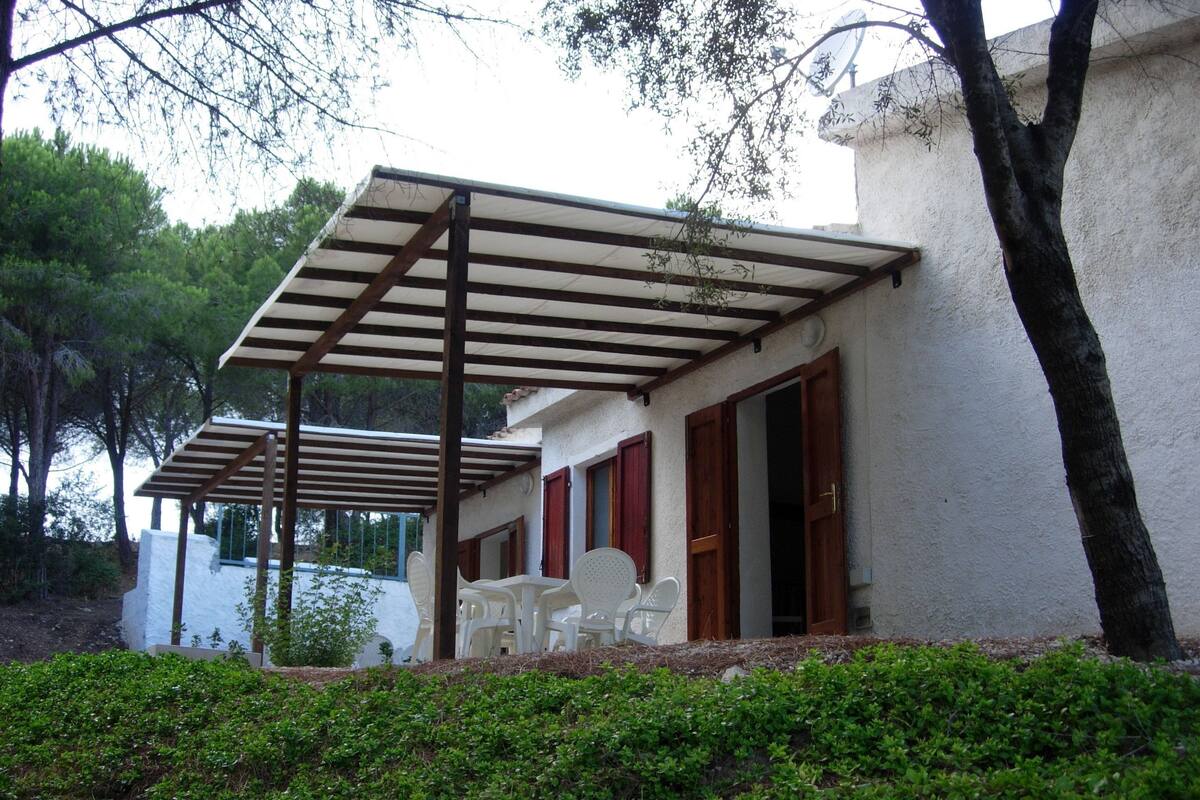 A covered outdoor seating area with white plastic chairs, a pergola, and a white building with red shutters.