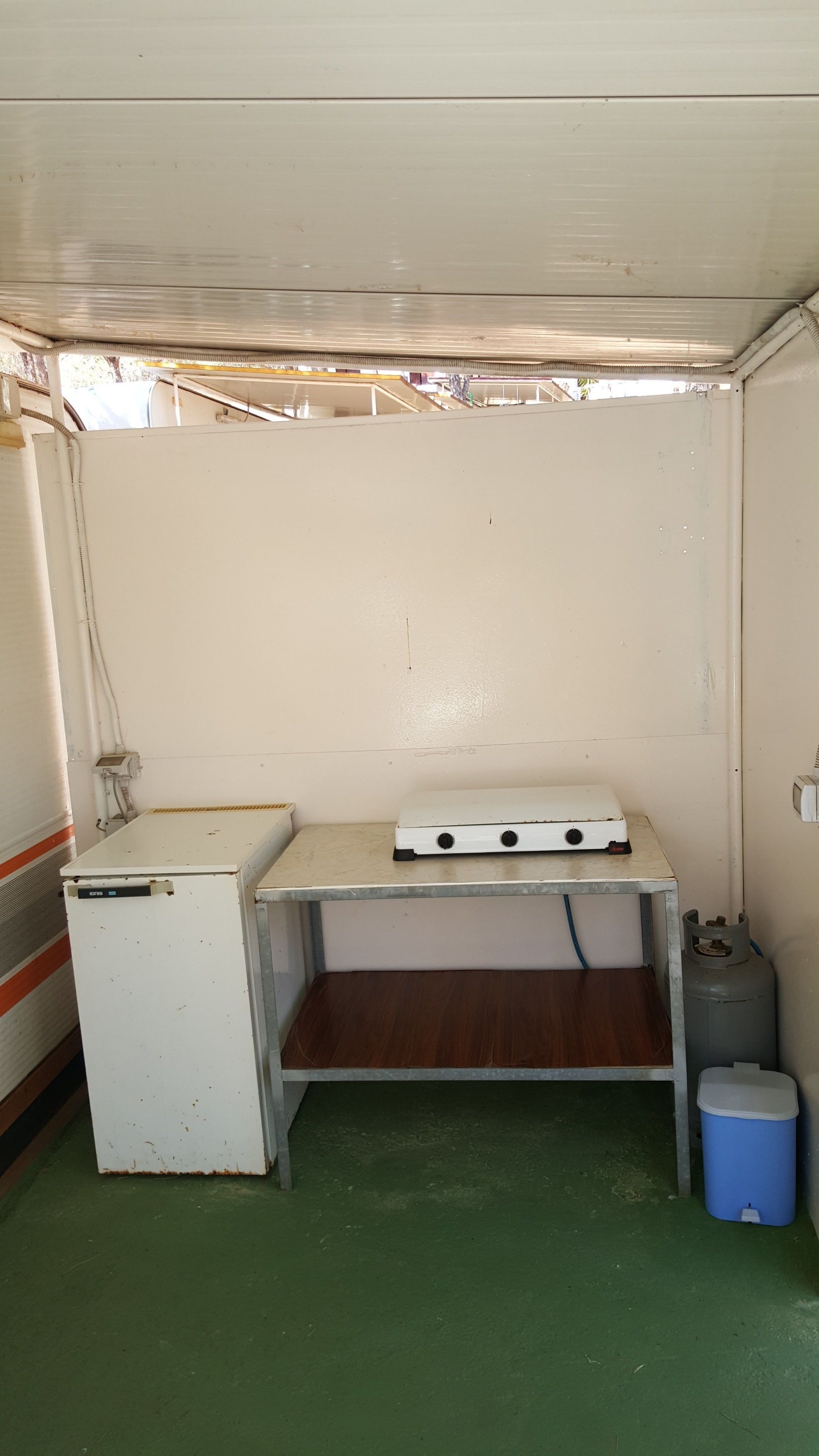 A compact room with a small kitchenette, a wooden shelf, a blue trash bin, and a green floor.