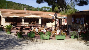 An outdoor café with tables and chairs, surrounded by potted plants and a tiled roof.