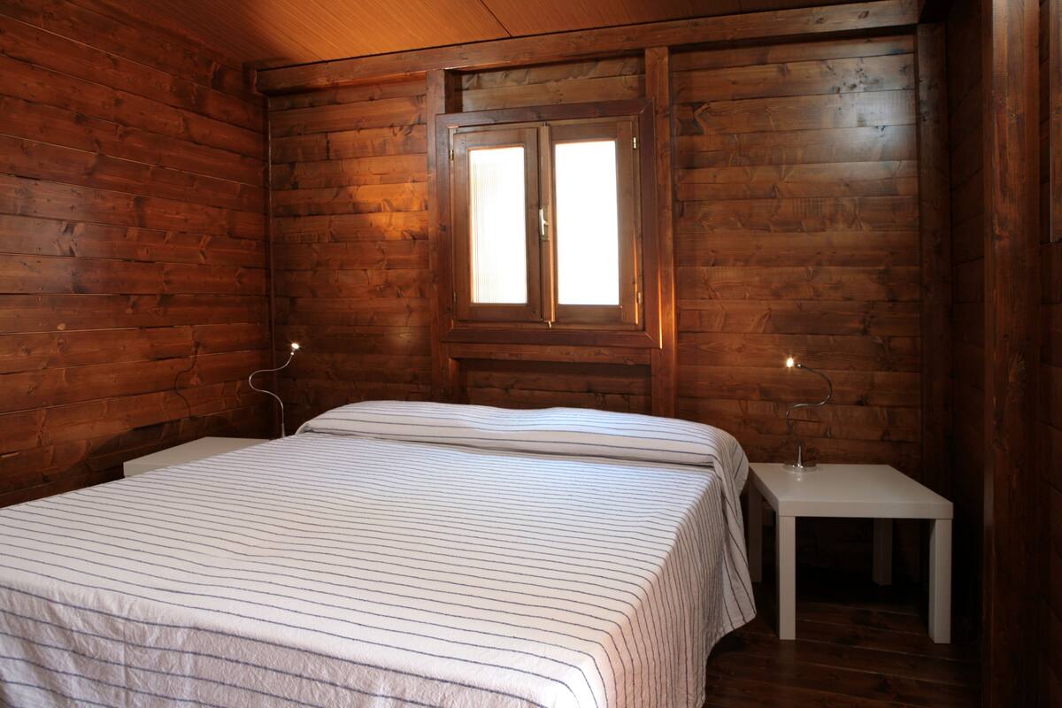 A wooden cabin room with a bed, a nightstand, and a window.