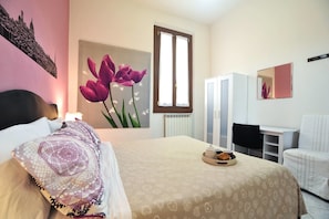 Hotelkamer met een bed, een bureau, een stoel, een radiator, een raam, een spiegel en een schilderij van tulpen.