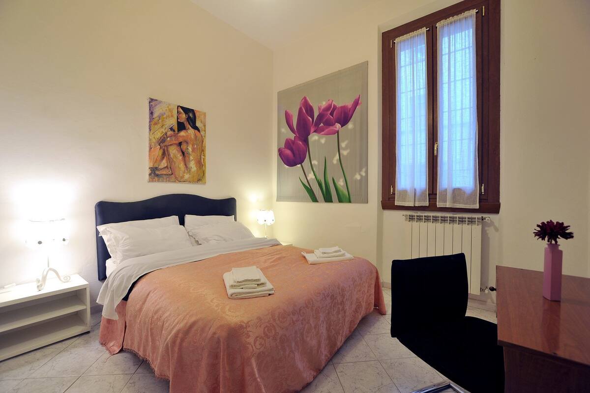 A neatly made bed with a pink bedspread and white pillows, a painting of a woman on the wall, a window with sheer curtains, and a vase with flowers on a wooden table.