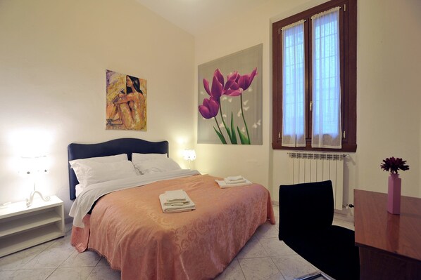 Una cama bien hecha con una colcha rosa y almohadas blancas, un cuadro de una mujer en la pared, una ventana con cortinas traslúcidas y un jarrón con flores sobre una mesa de madera.